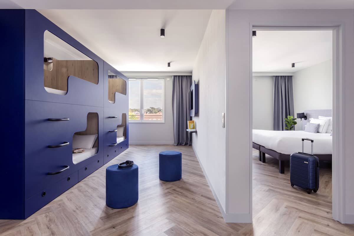 Una habitació d'hotel moderna amb un armari gran, dos otomans, un llit amb una maleta i un televisor penjat a la paret.