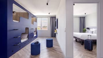 A modern hotel room with a large wardrobe, two ottomans, a bed with a suitcase, and a TV on the wall.