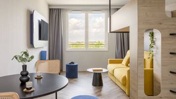 Uma sala de estar moderna com um sofá amarelo, uma mesa redonda com um vaso e porta-copos, um banquinho azul, um armário de madeira com uma planta e uma grande janela com vista para o campo.