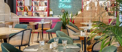 A cozy cafe with round tables and green chairs, a bookshelf in the background, and a neon sign that reads "The Kind Place".
