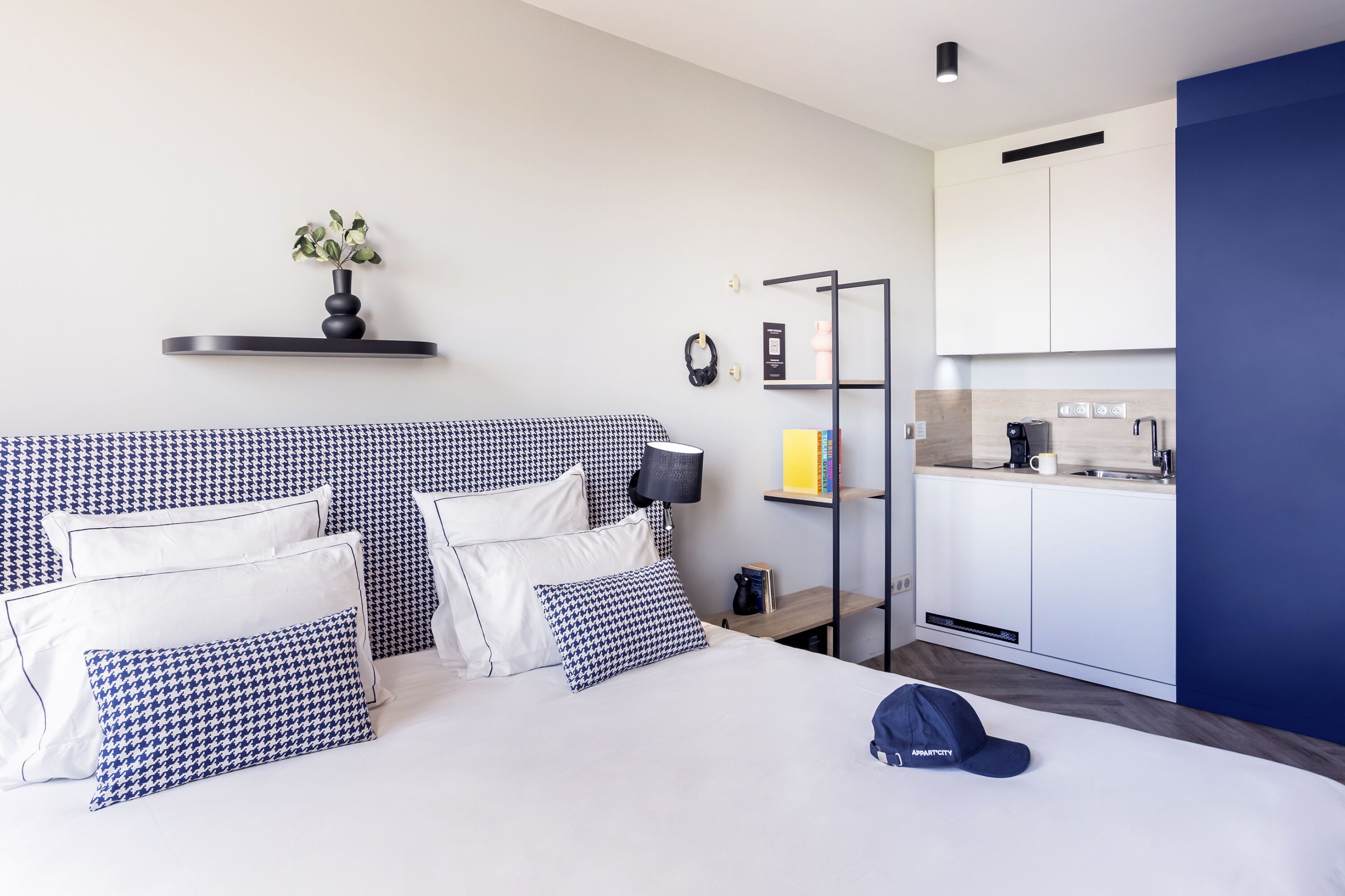 A hotel room with a bed, bedside table, lamp, and a small shelf with a plant.