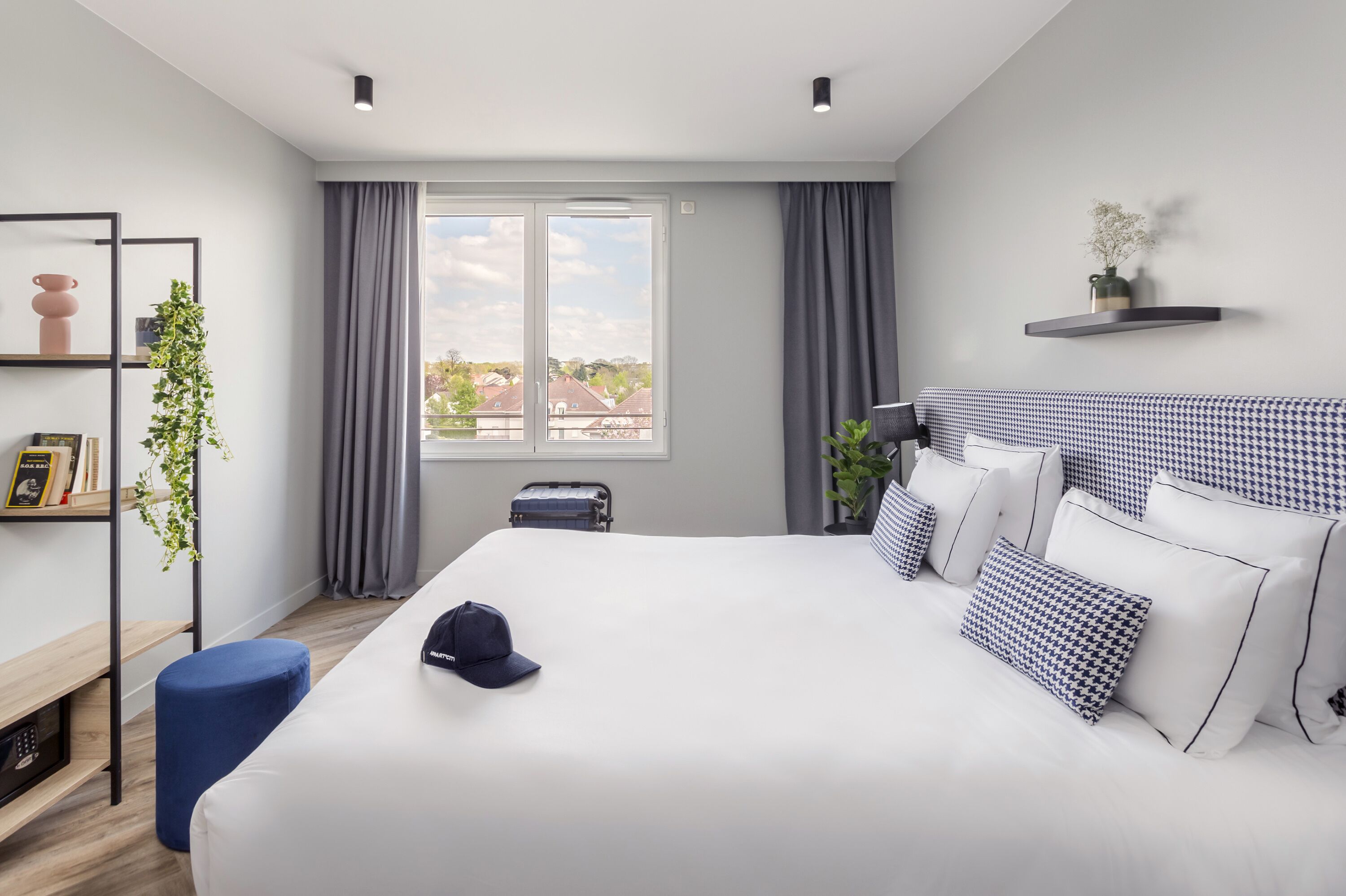 A modern hotel room with a large bed, a bedside table, a small desk, a bookshelf, and a window with curtains.
