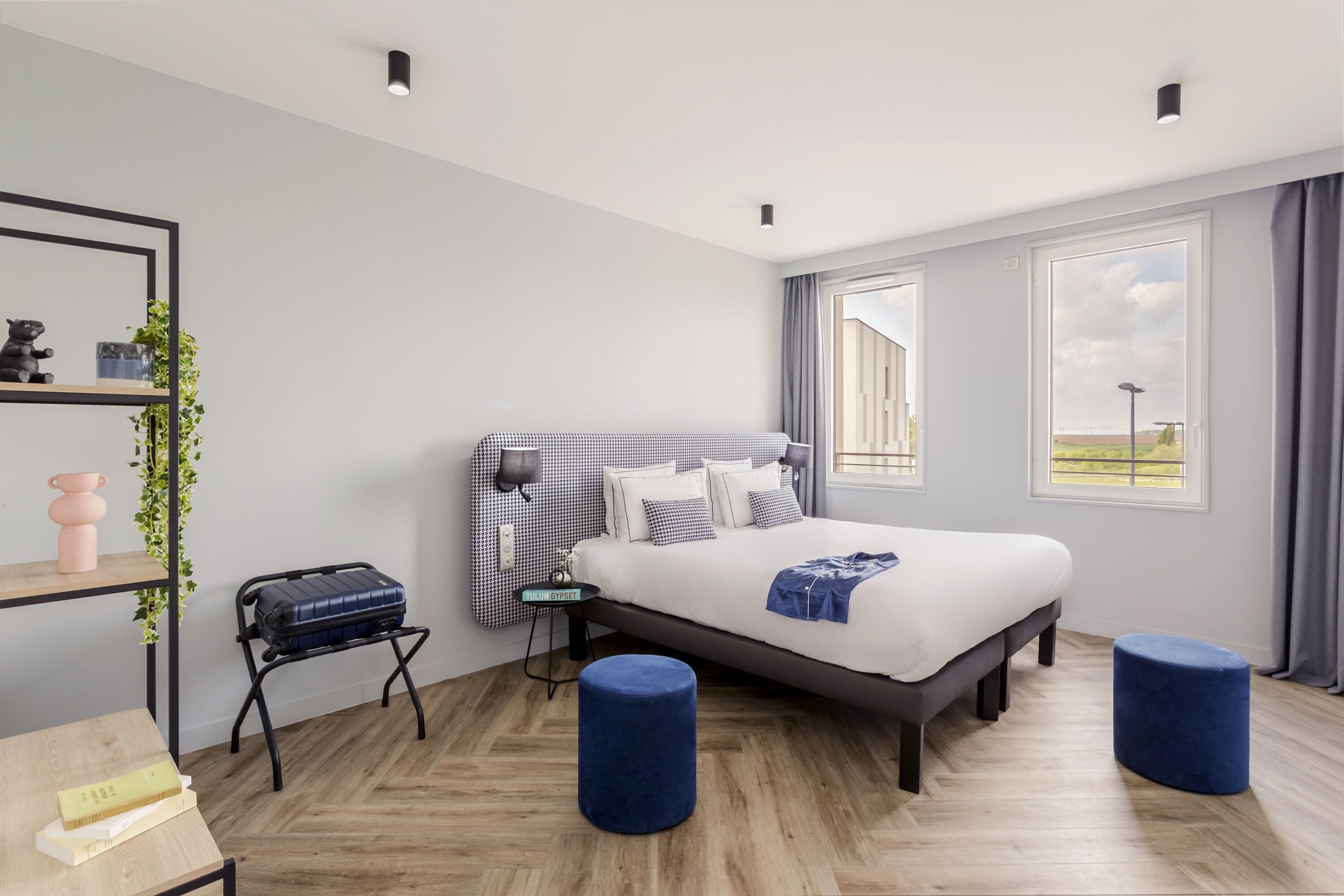 Quarto de hotel moderno com uma cama grande, uma escrivaninha com cadeira, uma mesa pequena e um banquinho azul.