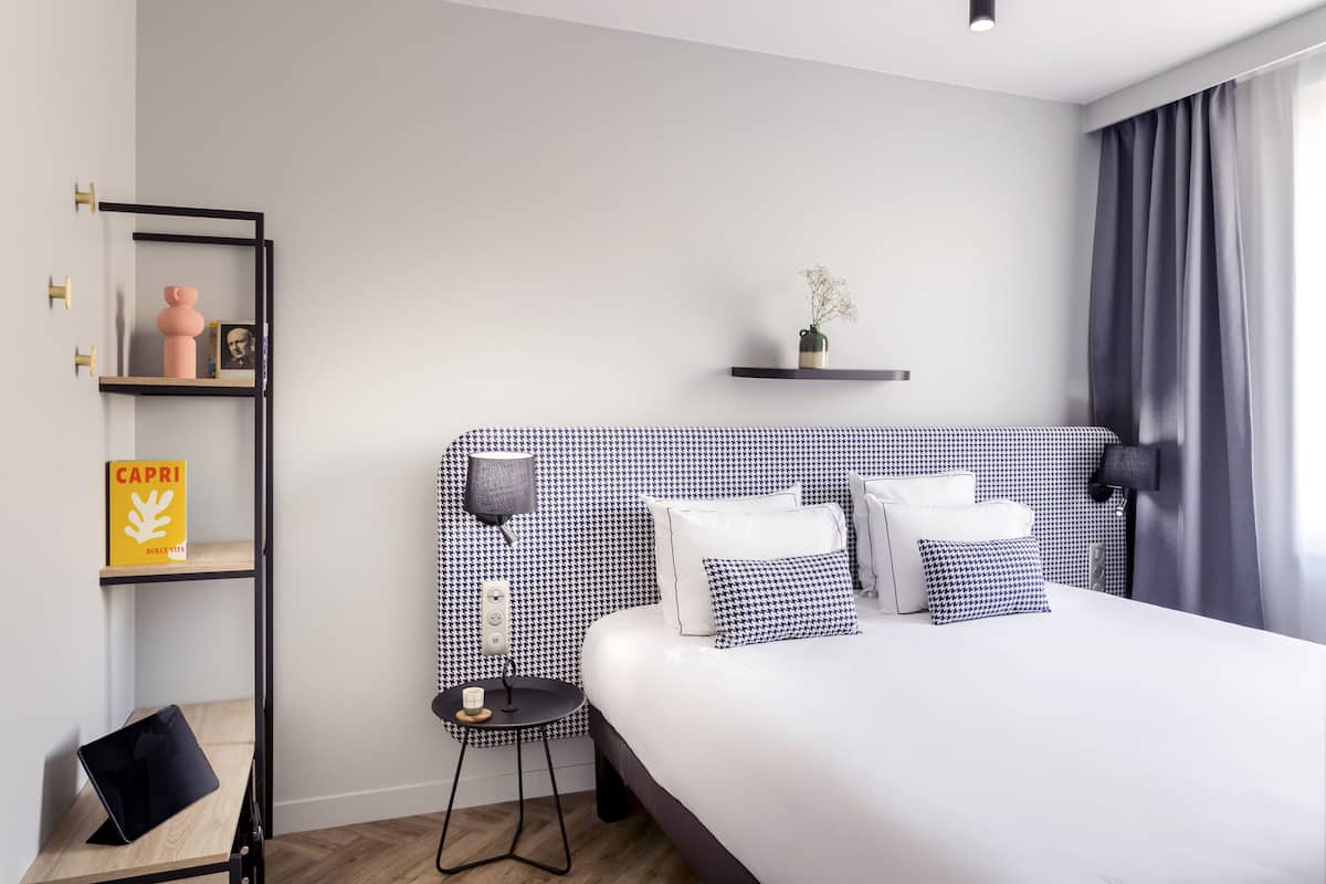 Habitació d'hotel moderna amb un llit gran, una taula de nit, una làmpada, una estant amb llibres i una petita planta.