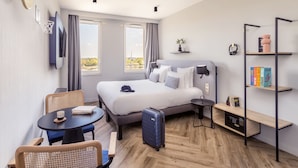 Una habitación de hotel moderna con una cama grande, un escritorio, una silla, una mesita, una estantería y una maleta.