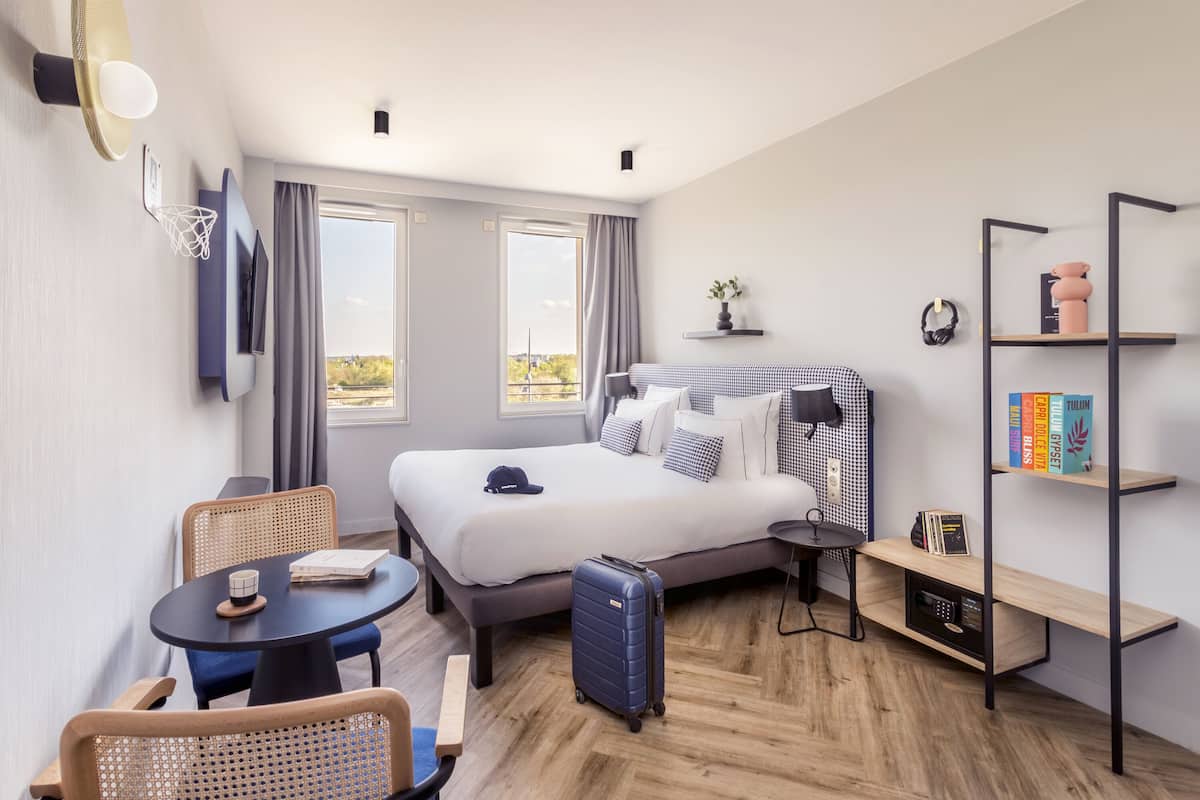 Habitació d'hotel moderna amb un llit gran, una taula de treball, una cadira, una taula petita, una prestatgeria i una maleta.
