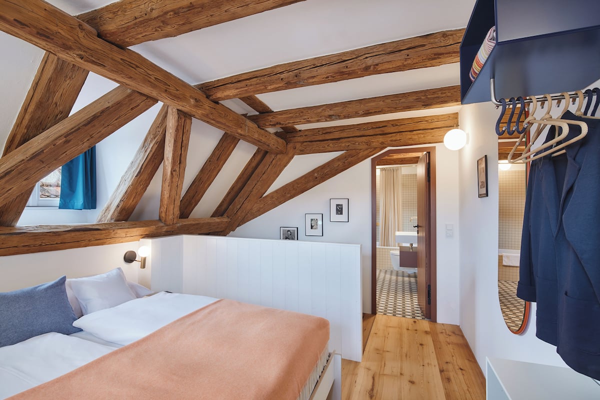 A bedroom with a sloped ceiling, wooden beams, a bed with a peach-colored blanket, a white headboard, and a bathroom visible through an open door.
