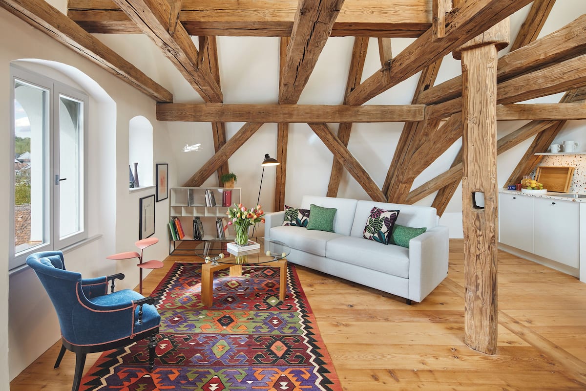 A spacious living room with exposed wooden beams, a white sofa, a blue armchair, and a colorful rug.