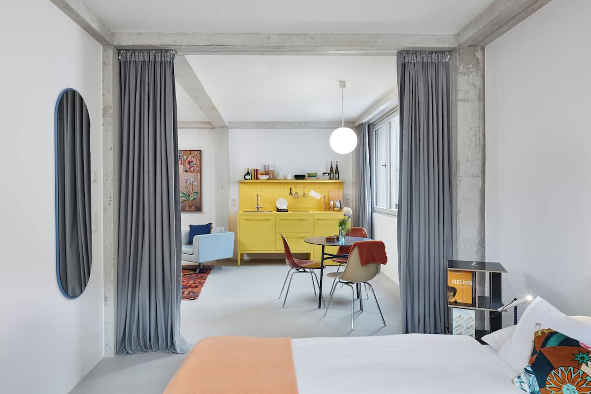 A modern hotel room with a large bed, a dining area with chairs, and a kitchen visible through an open door.