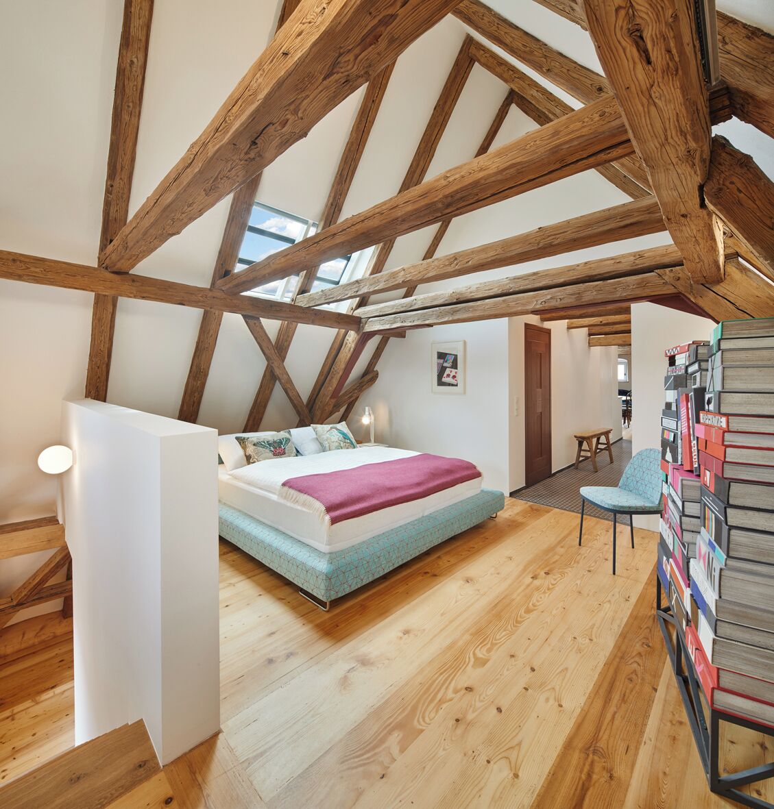 Un dormitorio con techo inclinado, vigas de madera, una cama, una silla y una estantería.