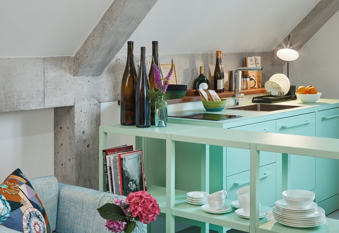 A kitchen with teal cabinetry, a countertop with wine bottles, a sink, and a dining area with white plates and bowls.
