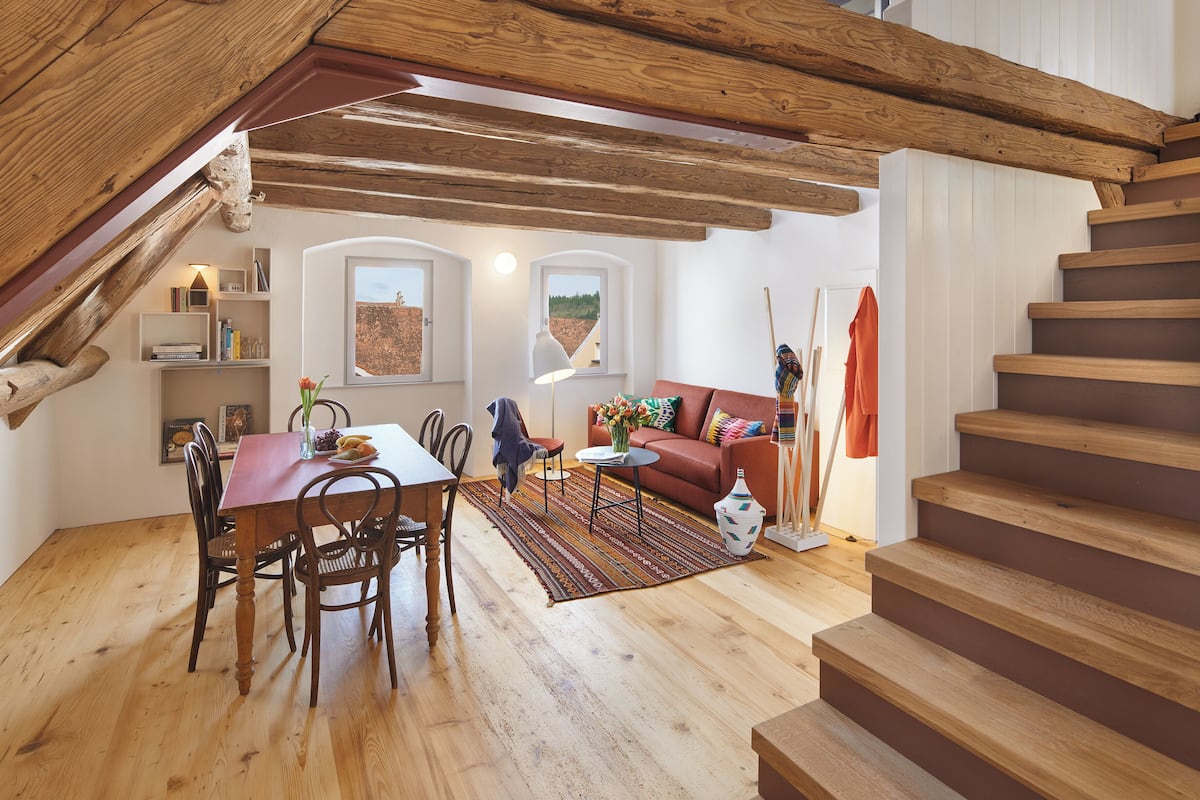 A spacious loft with exposed wooden beams, a dining area, a sofa, and a staircase.