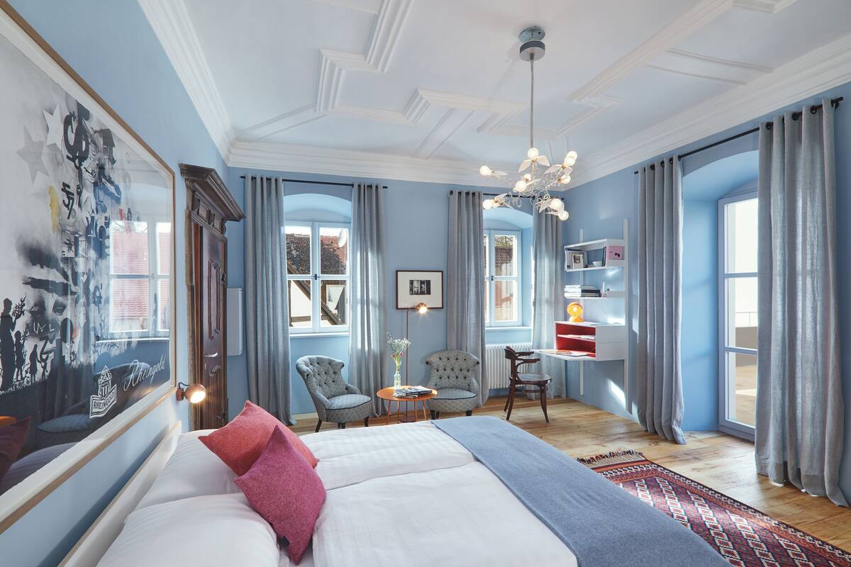 A spacious bedroom with a large bed, two armchairs, a small table, and a desk with a chair. There are large windows with curtains, a chandelier, and a wall-mounted shelf with decorative items.