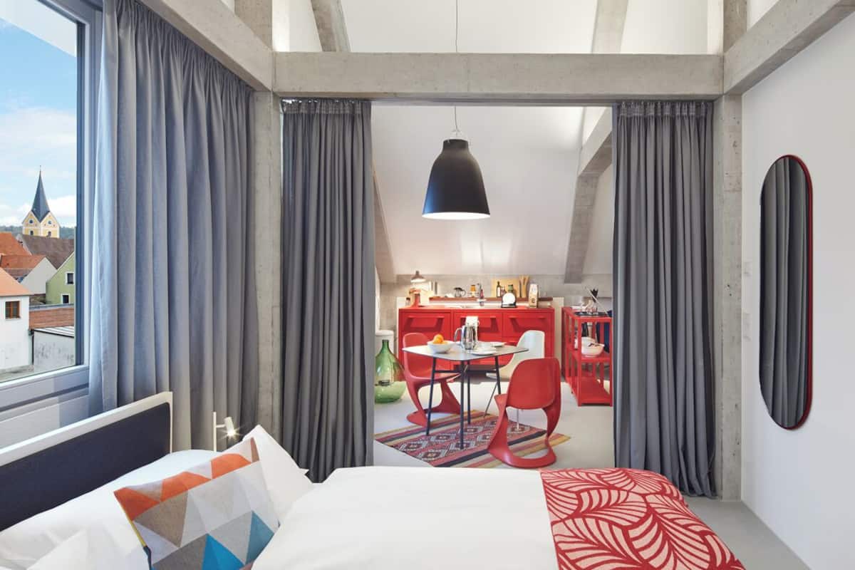 A modern hotel room with a large bed, a dining area, and a view of the city.