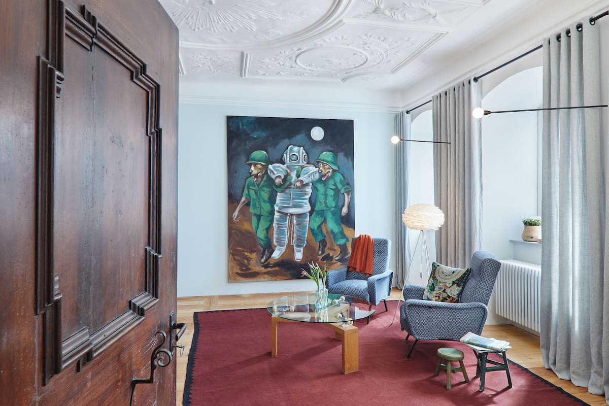 A living room with a large painting, a glass coffee table, a blue armchair, and a red rug.