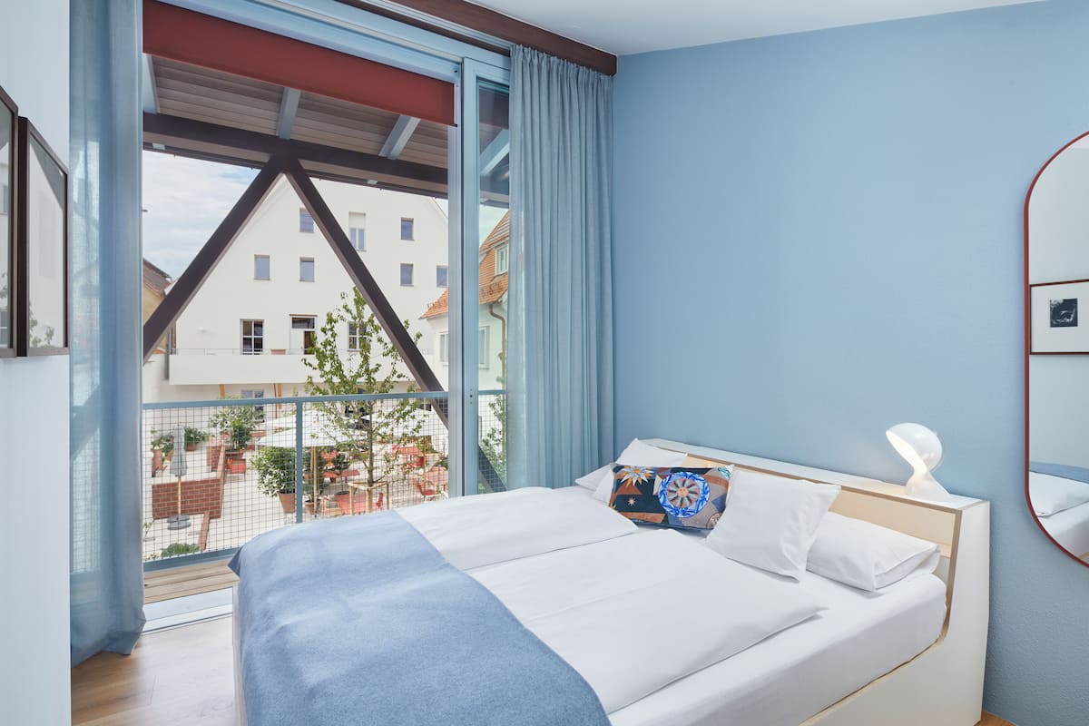 A bedroom with a bed, a window with curtains, a view of buildings outside, and a wooden floor.