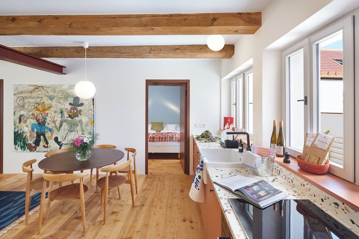 A cozy room with wooden floors, a dining area with a table and chairs, a bed with a colorful bedspread, a kitchen area with a sink and wine bottles, and large windows with a view of a building outside.