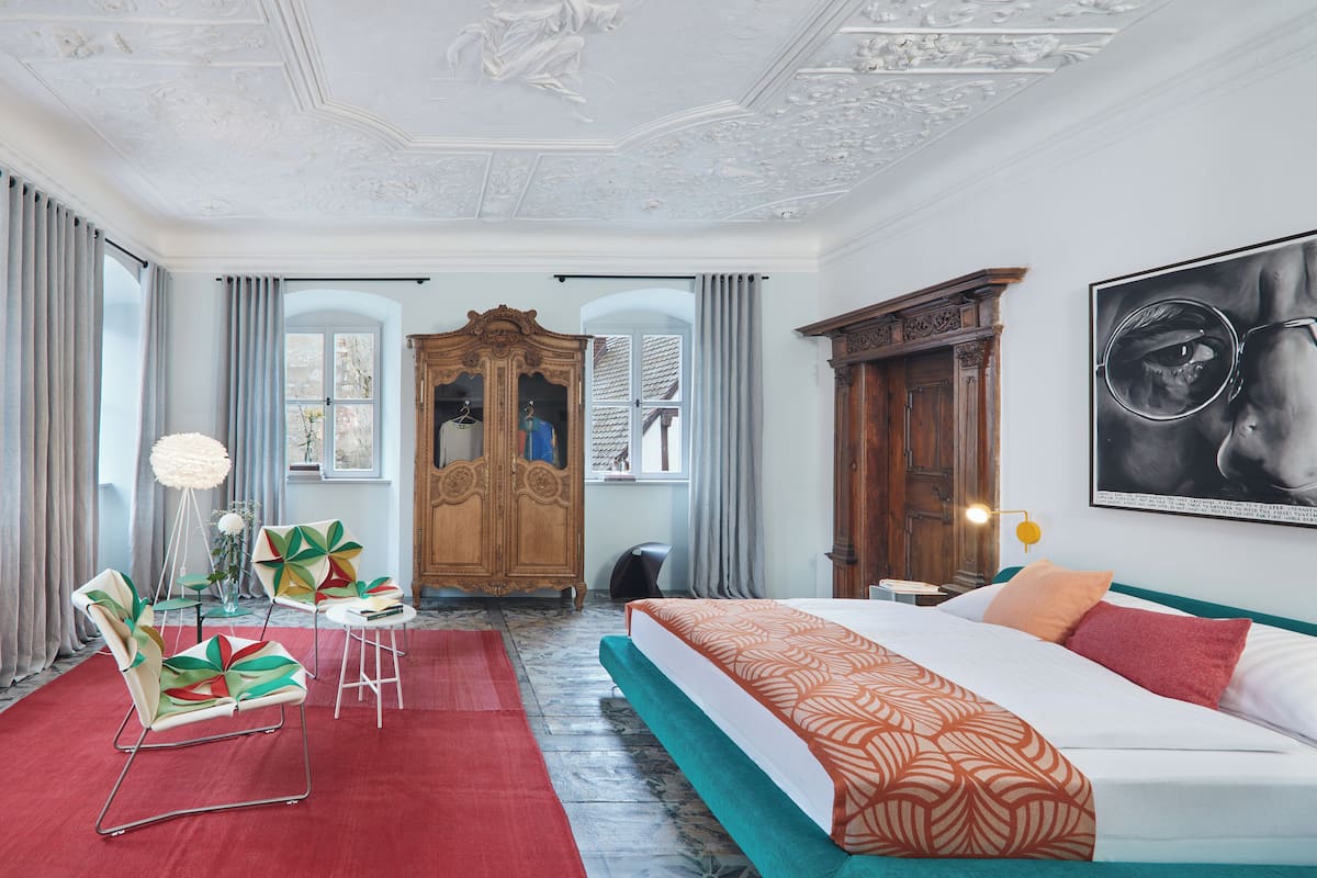 A hotel room with a large bed, a wooden wardrobe, a red carpet, and a large window with curtains.