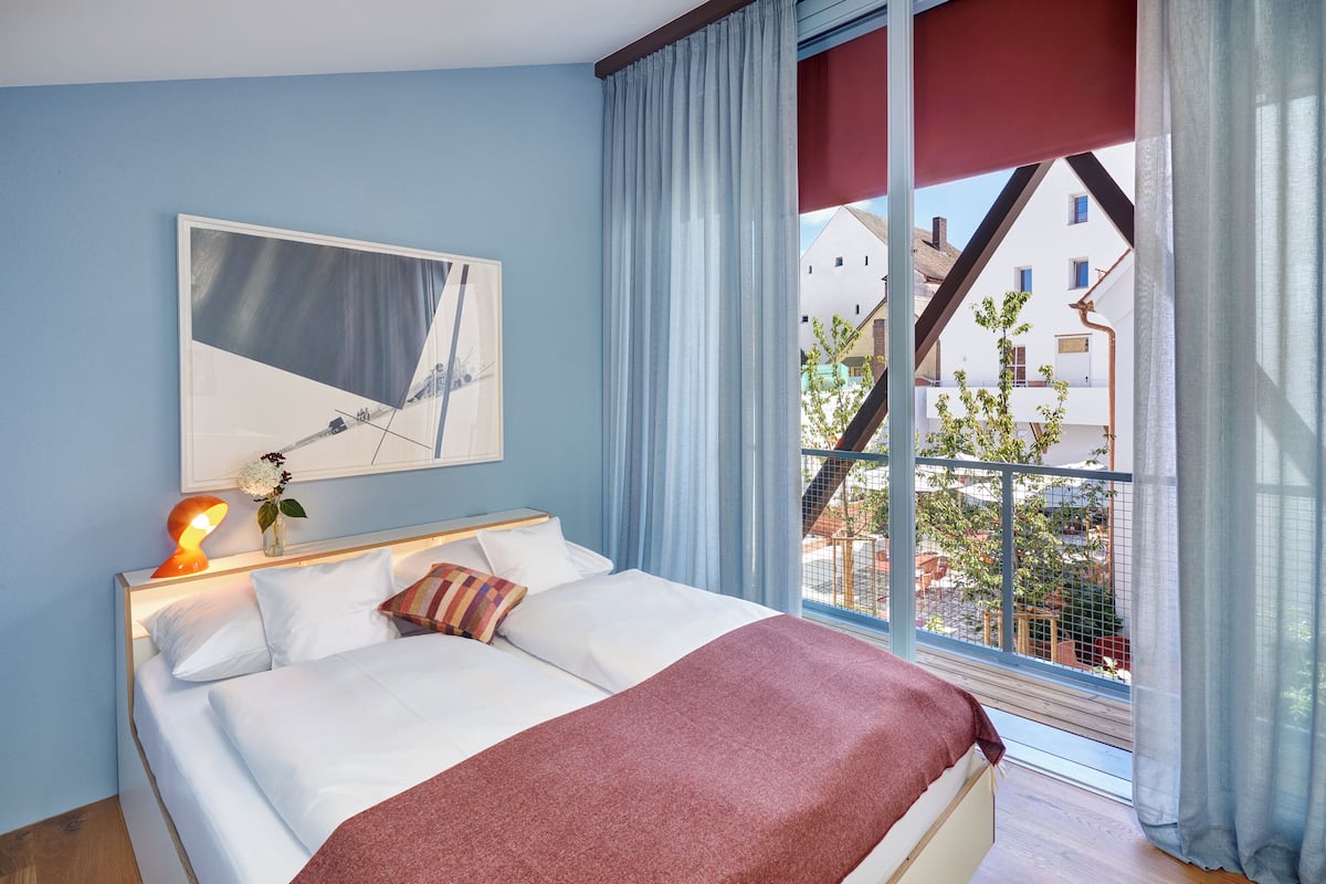A hotel room with a bed, a painting, a window with curtains, and a view of buildings outside.