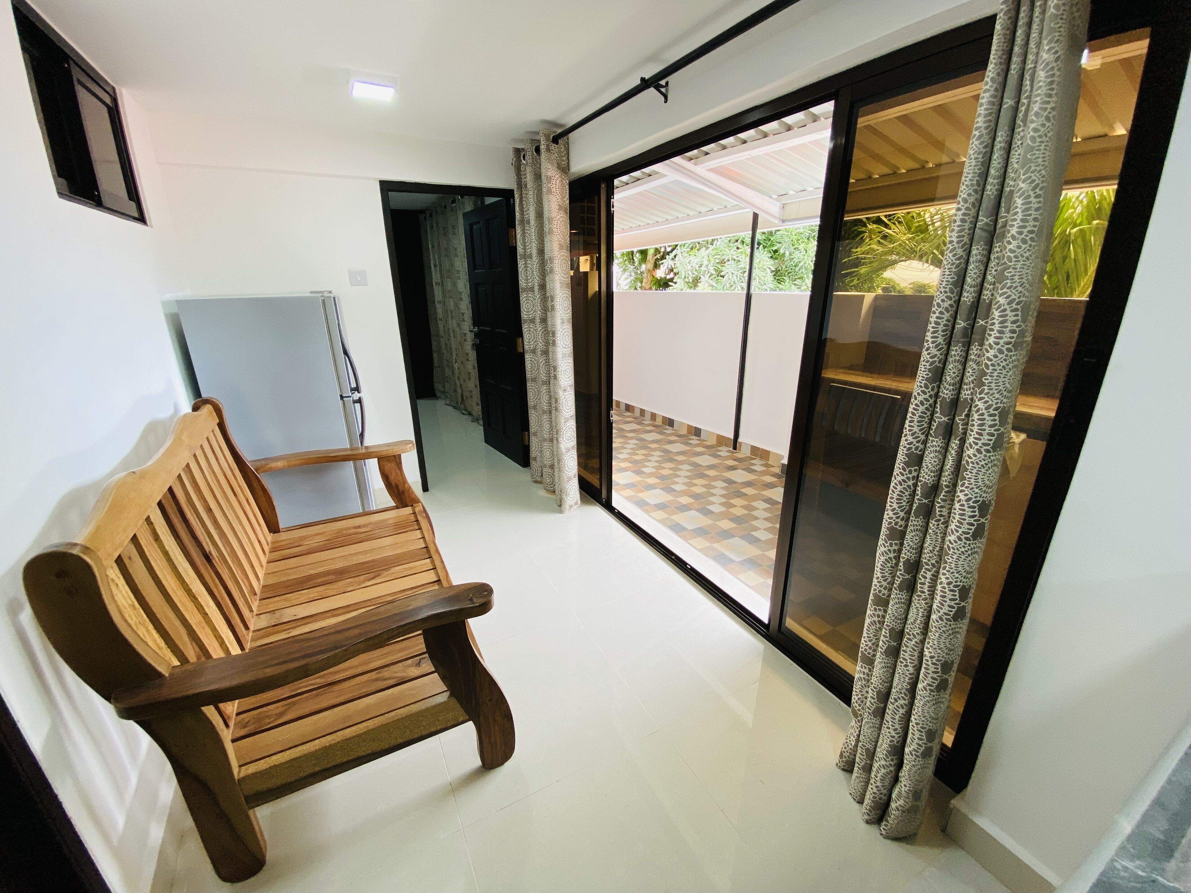 A room with a wooden bench, a refrigerator, a sliding glass door leading to an outdoor area with a tiled floor and a covered patio.