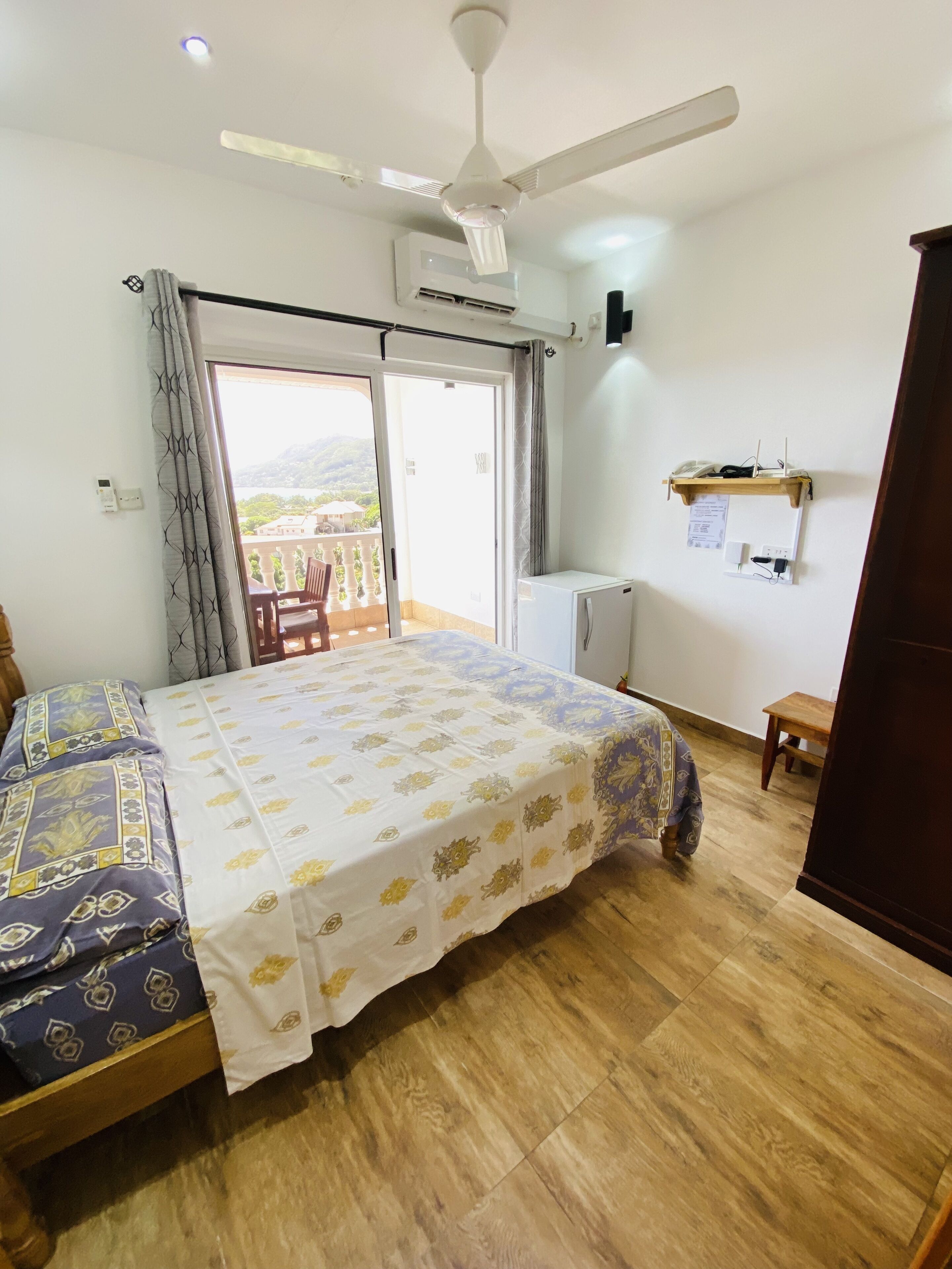 Un dormitorio con cama, ventilador de techo, equipo de aire acondicionado, piso de madera, balcón con vistas y una mesita.