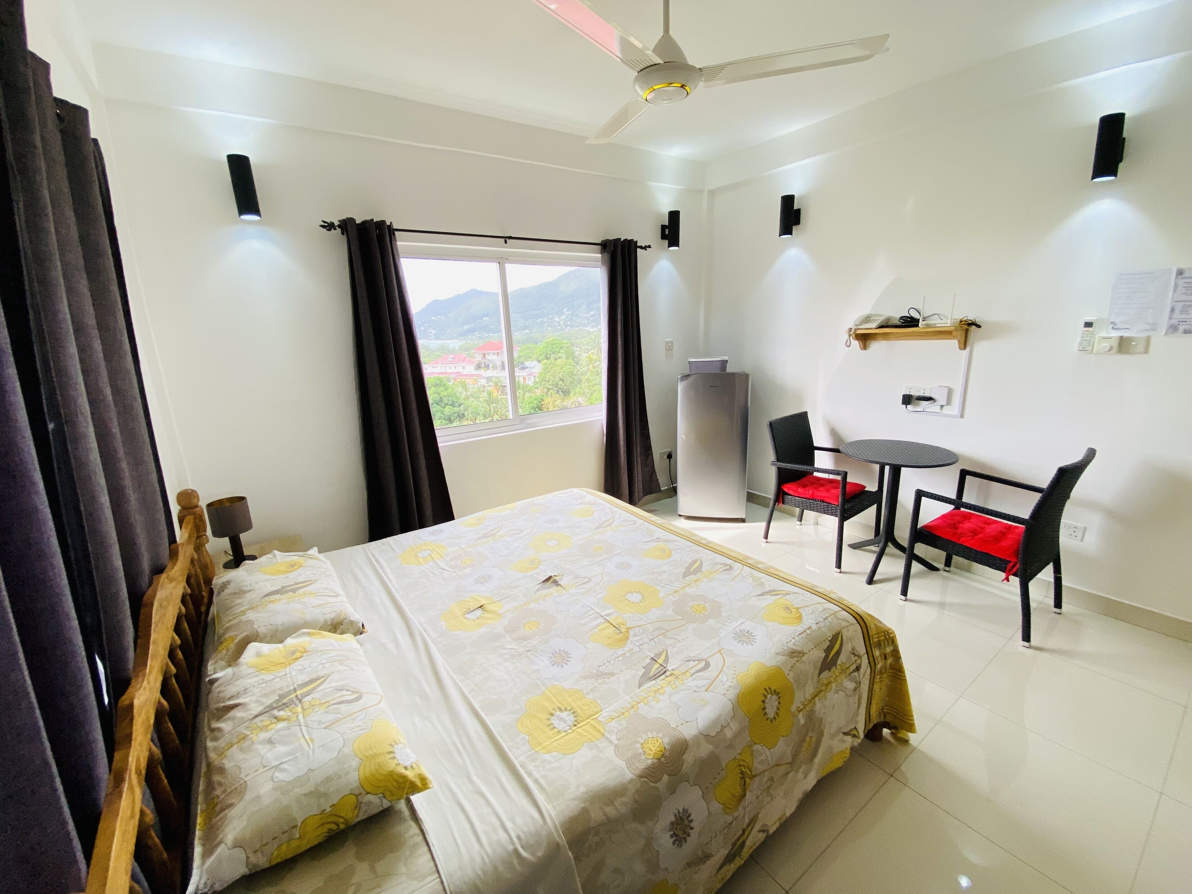 Une chambre d’hôtel comprenant un lit, un bureau avec deux chaises, un réfrigérateur et une fenêtre avec des rideaux.