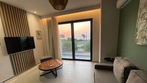 Un soggiorno moderno con una TV a schermo piatto, un tavolo rotondo in legno, un divano e una porta scorrevole in vetro che dà su un balcone con vista sul tramonto.