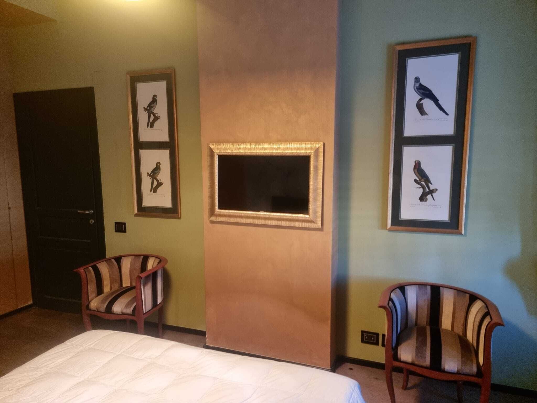 Quarto de hotel com uma cama, duas poltronas listradas, uma parede com ilustrações de pássaros emolduradas e uma porta.