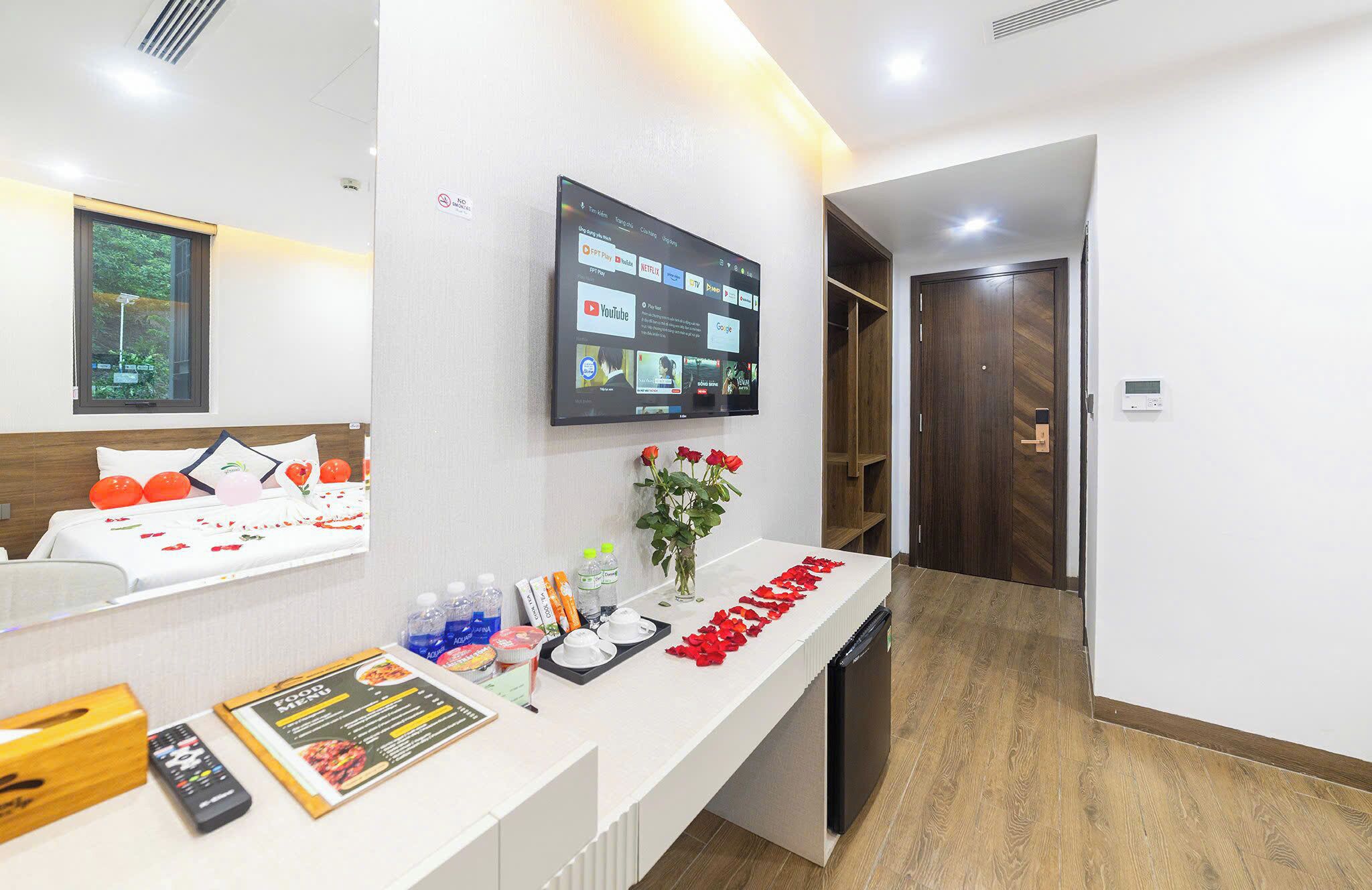 A hotel room with a bed, a TV mounted on the wall displaying various channels, a small table with a menu, and a wooden door.