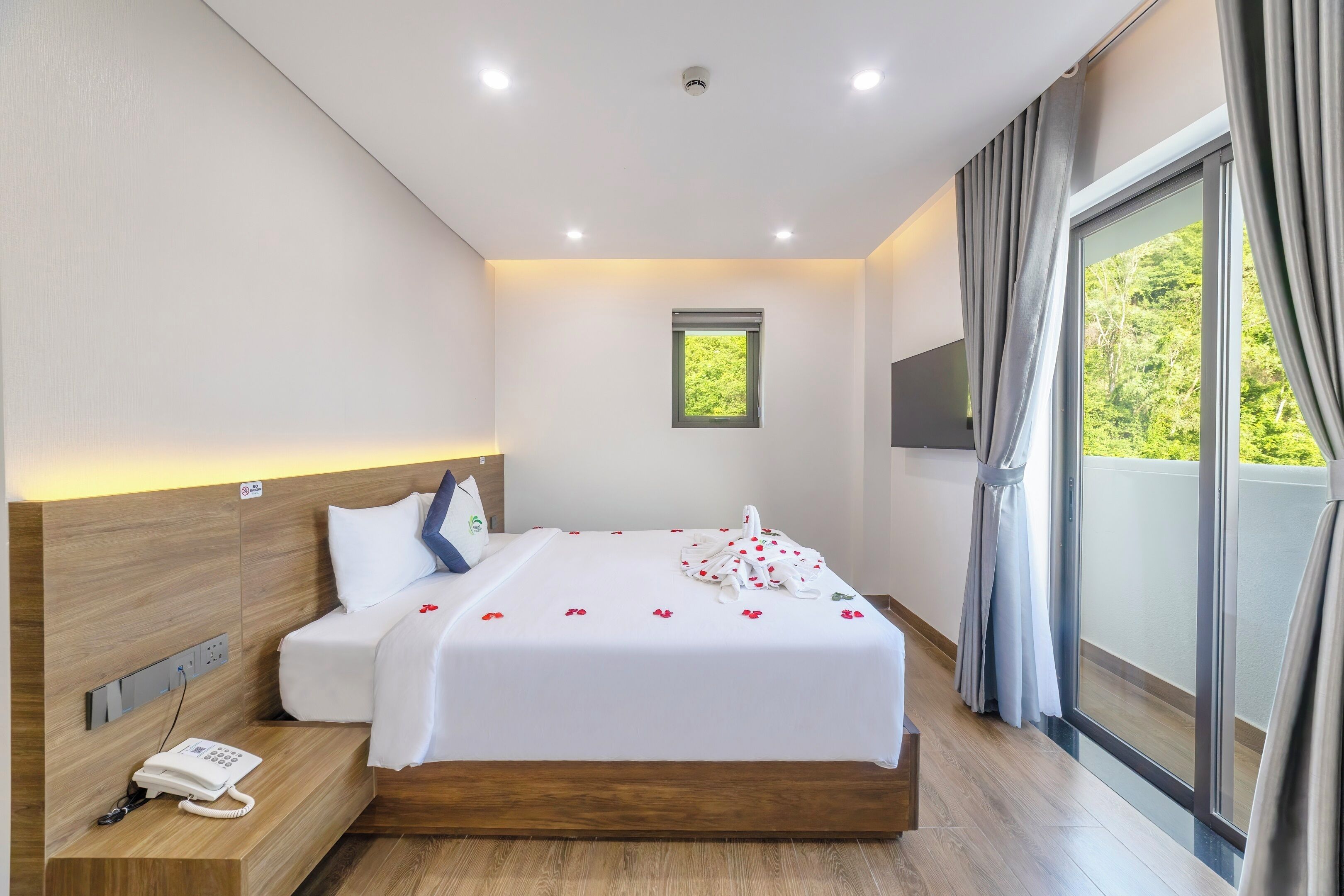 Una habitación de hotel moderna con cabecera de madera, una cama con ropa de cama blanca, un teléfono en una mesita de noche, un ventanal pequeño y vistas a zonas verdes a través de las cortinas.