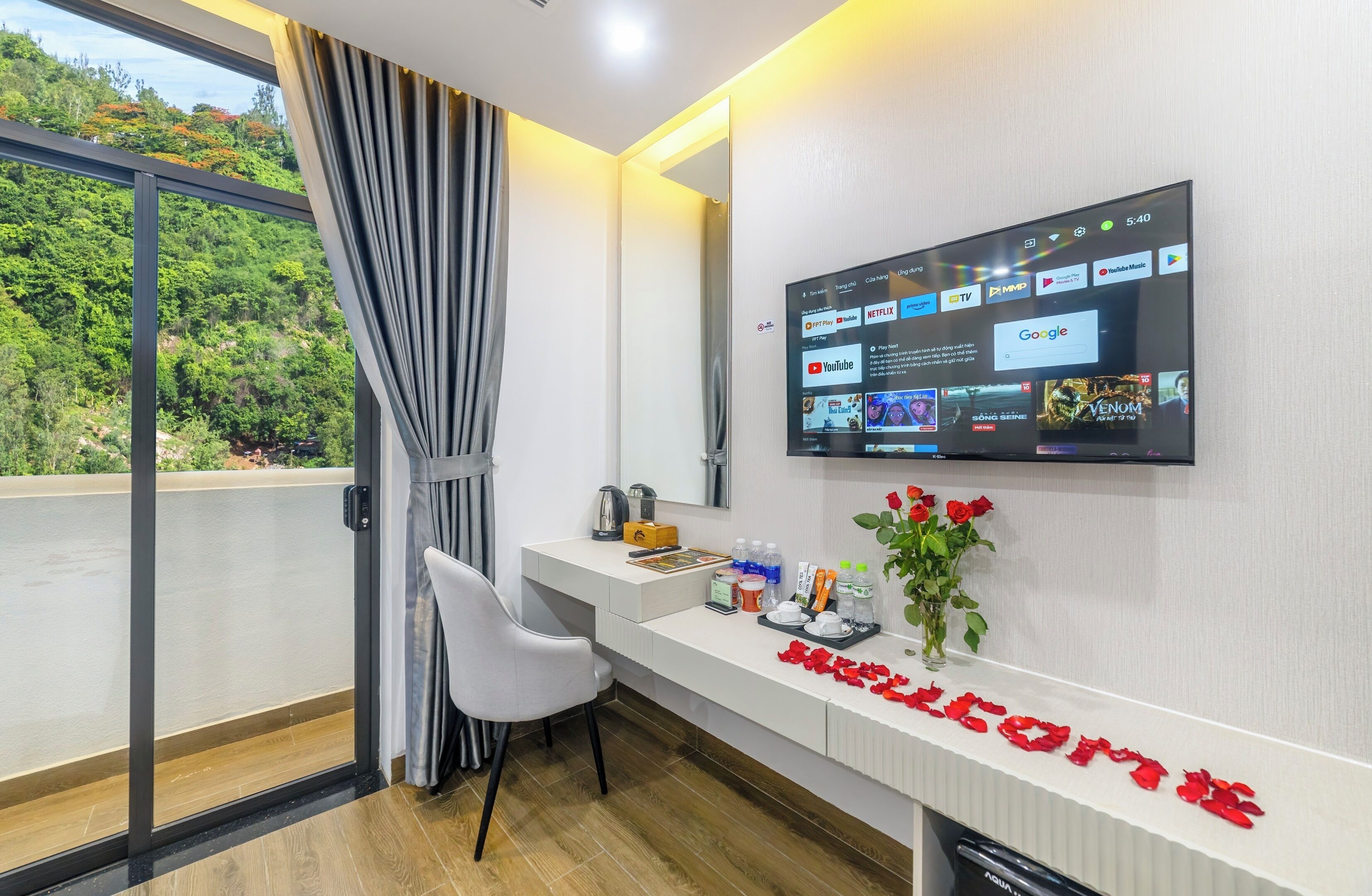 A modern hotel room with a large TV displaying various streaming services, a desk with a laptop, a chair, and a welcome sign with red roses.