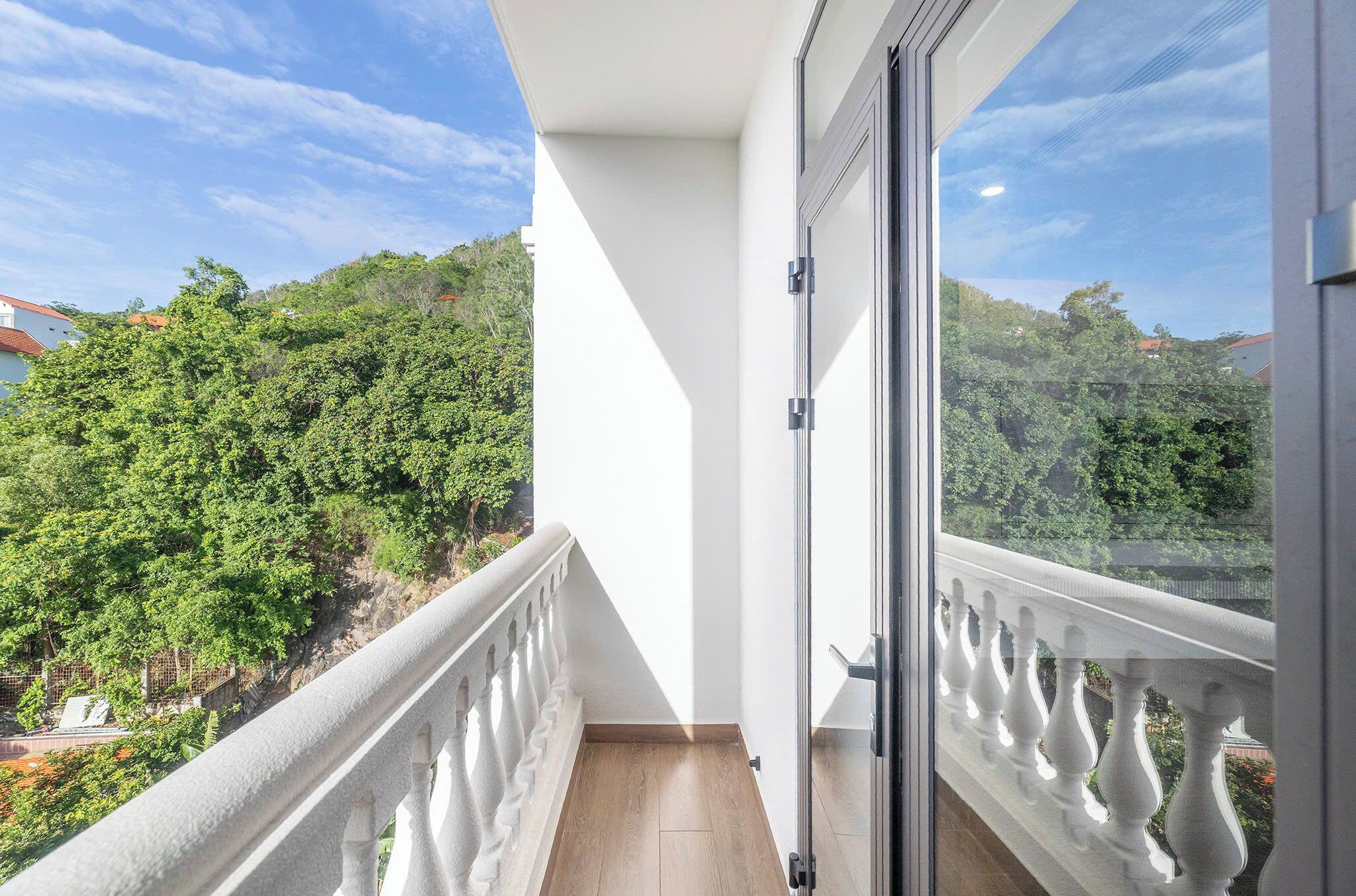 Un balcón con barandilla blanca, puerta de cristal y vista a un paisaje verde con árboles y cielo despejado.