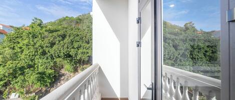 Un balcón con barandilla blanca, puerta de cristal y vista a un paisaje verde con árboles y cielo despejado.