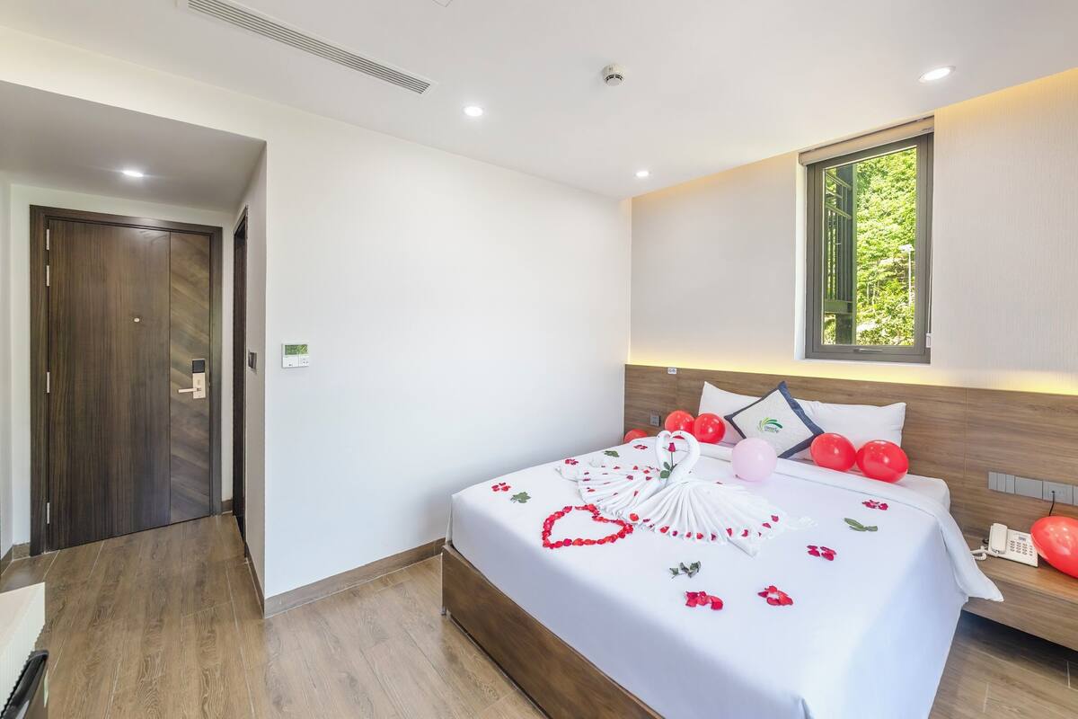 Una habitación de hotel con piso de madera, cama con cubrecama blanco, decoración con forma de corazón y ventana con vista a follaje.