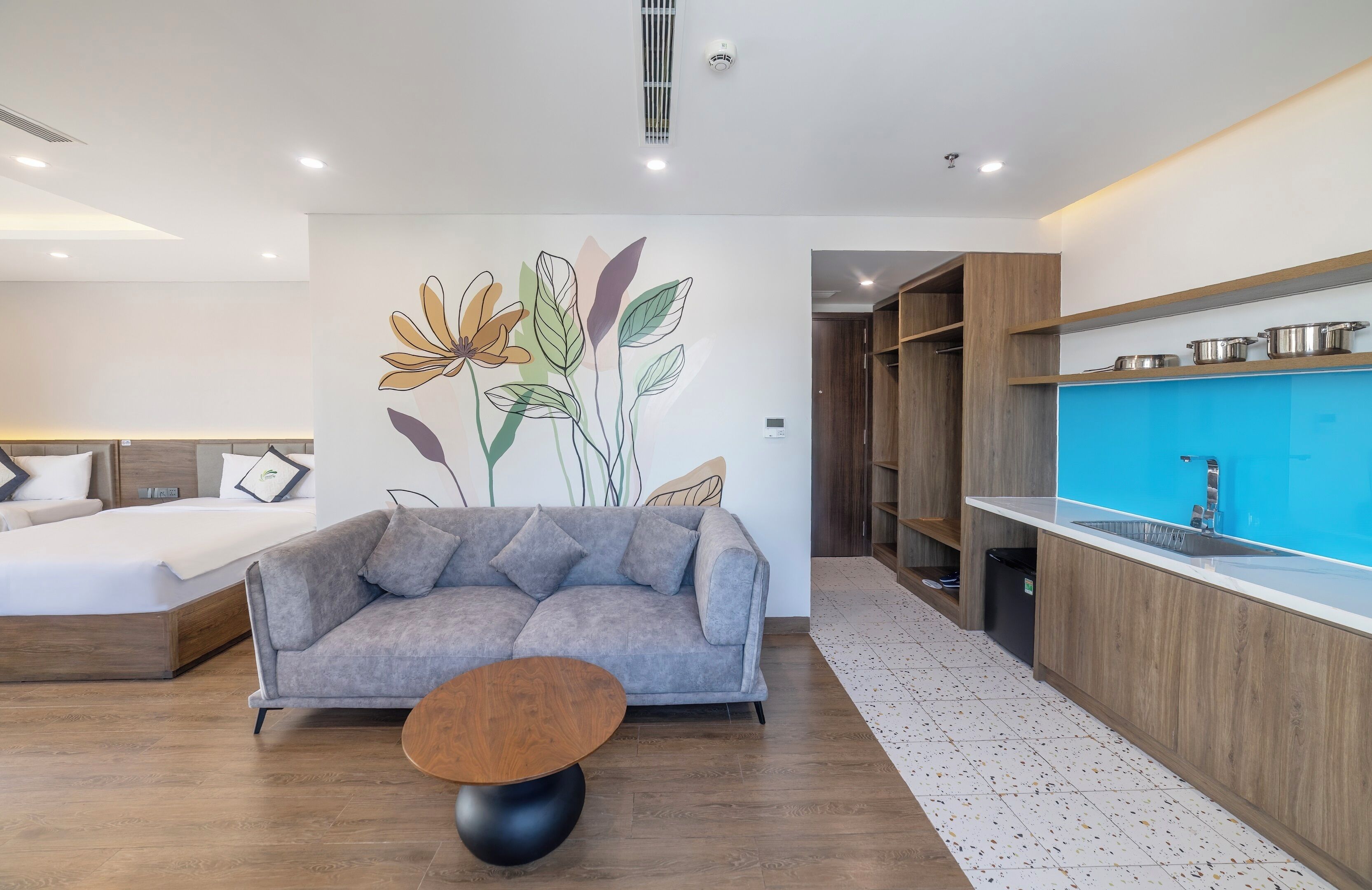 Una habitación moderna con un sofá gris, una mesita de centro redonda de madera, una kitchenette con encimera azul y una pared con un mural floral.