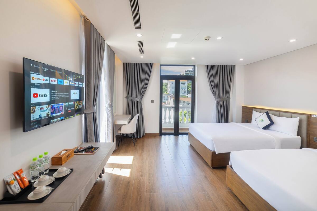 Habitación de hotel con dos camas, un escritorio, un televisor montado en la pared y vistas al exterior a través de una puerta de cristal.