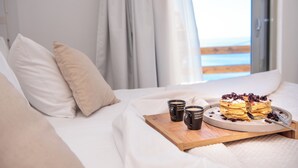 Un lit bien fait, avec du linge de lit blanc et des coussins beige, un plateau en bois sur lequel reposent deux tasses de café et une part de gâteau, et une vue sur la mer par une fenêtre ornée de rideaux.