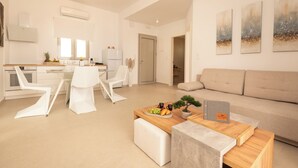 Un salon moderne comprenant un canapé, une table basse avec des fruits et un livre, un coin repas avec des chaises et une table, et une cuisine équipée d’appareils électroménagers.