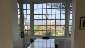 A dining area with a table and chairs, large windows with a view of greenery and buildings, and a hanging light fixture. - Maison vue mer à Bénerville (bénerville sur mer)