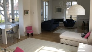 A spacious living area with a white sofa, a blue sofa, a pink chair, and a white dining table with chairs. There is a large window providing natural light and a view of the outdoors. - Maison vue mer à Bénerville (bénerville sur mer)