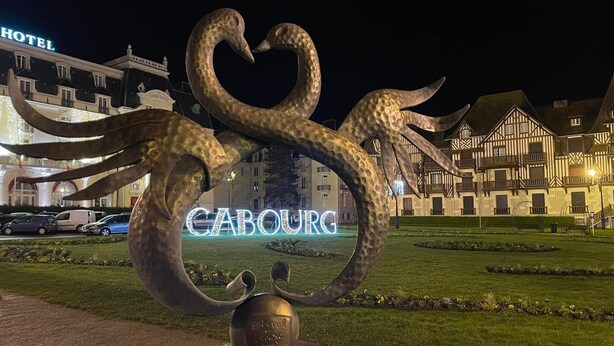 Une sculpture représentant des serpents entrelacés, avec une plaque à sa base. En arrière-plan, on aperçoit un hôtel avec une enseigne indiquant « HÔTEL », et un bâtiment sur lequel le nom « CABOURG » est illuminé au premier plan.