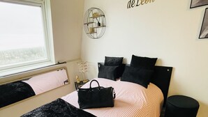 Une chambre à coucher avec un lit, une literie noire et rose, un sac à main noir posé sur le lit, une petite table ronde avec une lampe, et un mur orné d’une étagère circulaire et d’un texte.