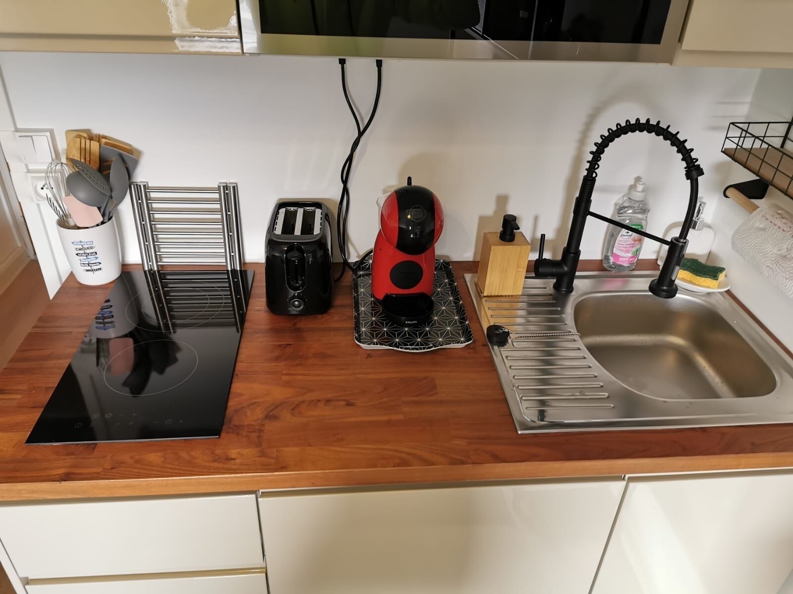 A kitchen with a black induction cooktop, a toaster, a red coffee maker, a sink with a faucet, and a wooden cutting board.
