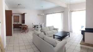 A spacious living room with a sofa, a dining table with chairs, a kitchen area, and large windows allowing natural light. - Anavyssos Dream Villa Sea View and Pool (Saronikos)