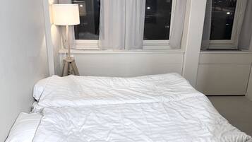 A bedroom with a bed, a nightstand with a lamp, and two windows with curtains.