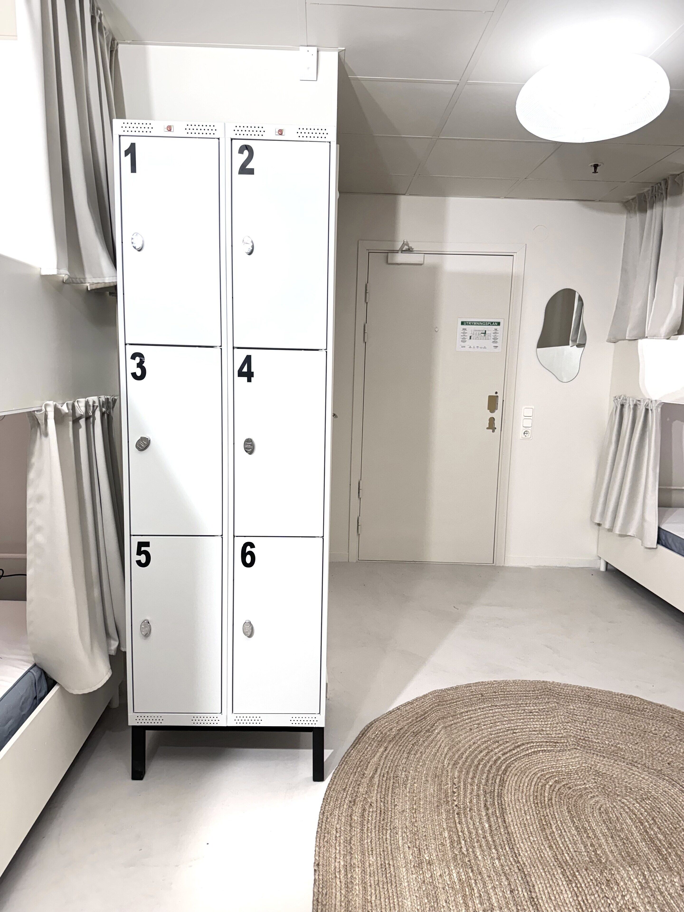 Une chambre d’hôtel dotée d’une grande armoire numérotée de 1 à 6, d’un lit avec une tête de lit, d’un tapis rond en fibres tressées et d’une porte avec une pancarte.