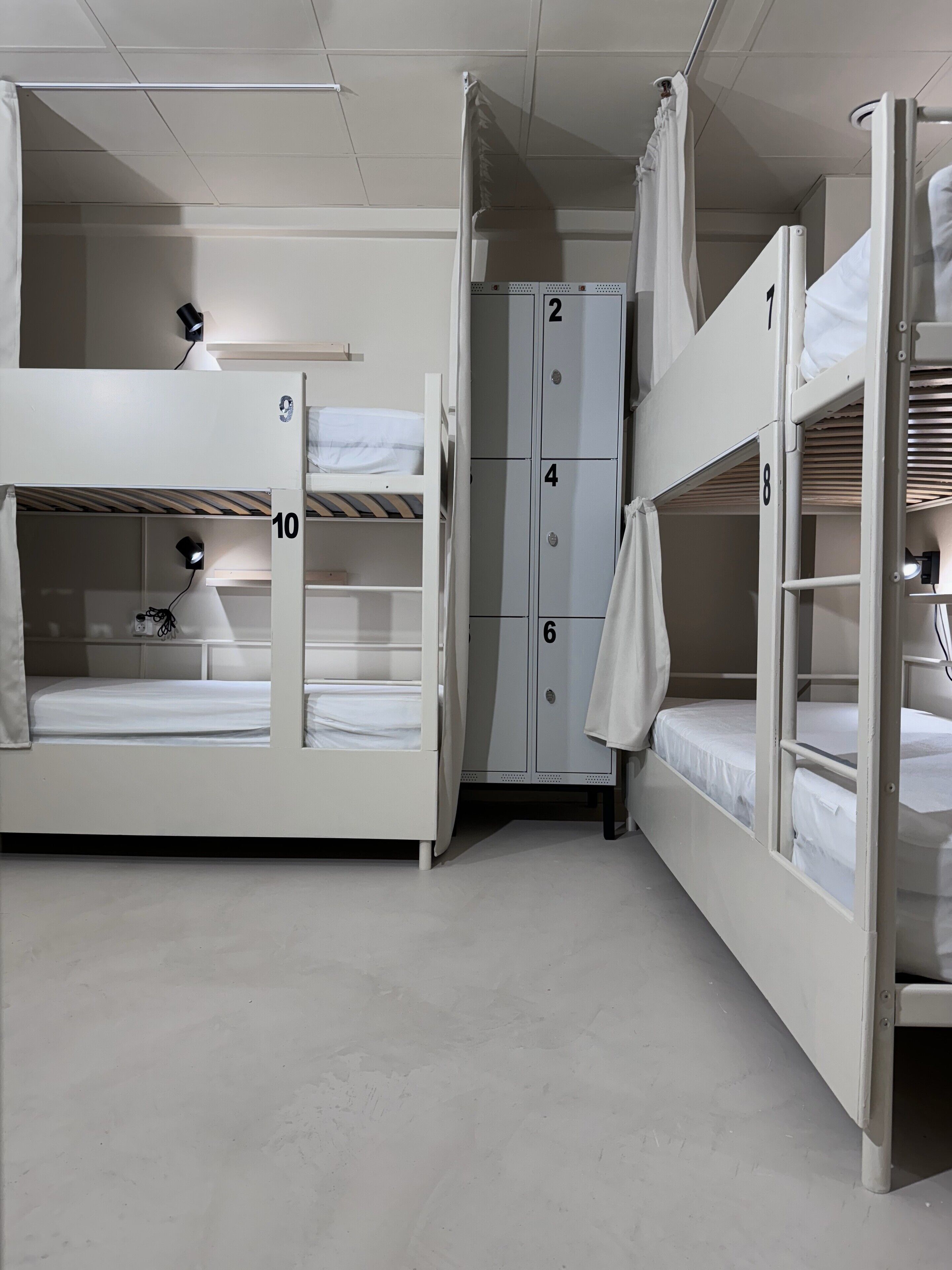 Une chambre de type dortoir, avec des lits superposés, des casiers numérotés et un bureau équipé d’une lampe.