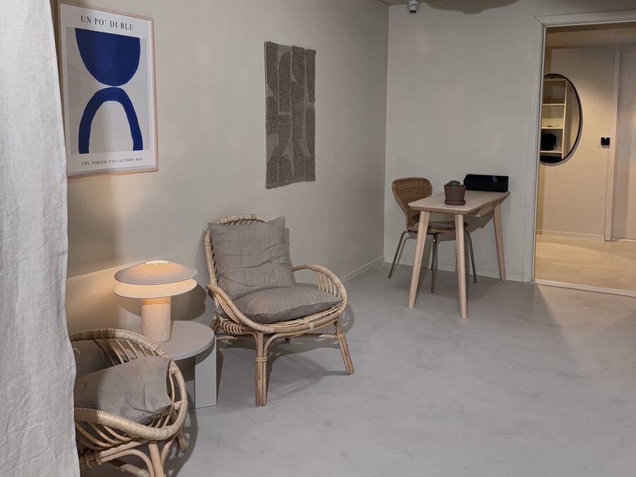 A modern, minimalist room with a white floor and walls, featuring a wicker chair, a small round table, and a wall-mounted lamp.