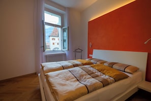 A bedroom with two beds, a window with a view of buildings, and a red accent wall.