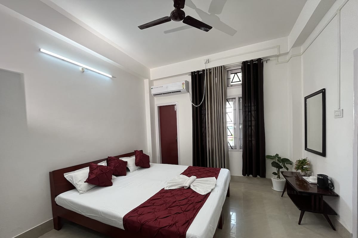 A hotel room with a bed, a ceiling fan, a desk with a chair, a potted plant, and a window with curtains.